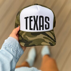 A person's hand holding a camouflage baseball cap with the word "TEXAS" printed on it.