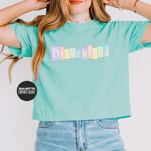 A woman wearing a mint green t-shirt with the word "Disneyland" printed on it, posing with her hands behind her head.
