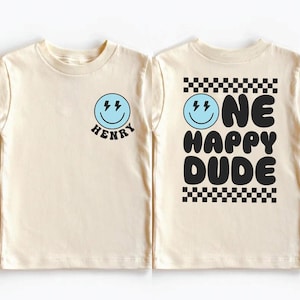 two white t-shirts with black text and graphics. The t-shirt on the left has a smiley face and the name "HENRY" below it, while the t-shirt on the right has the text "ONE HAPPY DUDE" and a checkered pattern.