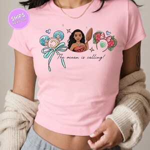 A woman wearing a pink t-shirt with a graphic design featuring a cartoon character and the text "The ocean is calling!"