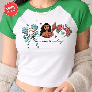A white and green t-shirt with a graphic design featuring a woman surrounded by various ocean-themed elements, including a surfboard, flowers, and a ribbon. The text on the t-shirt reads "The ocean is calling!"