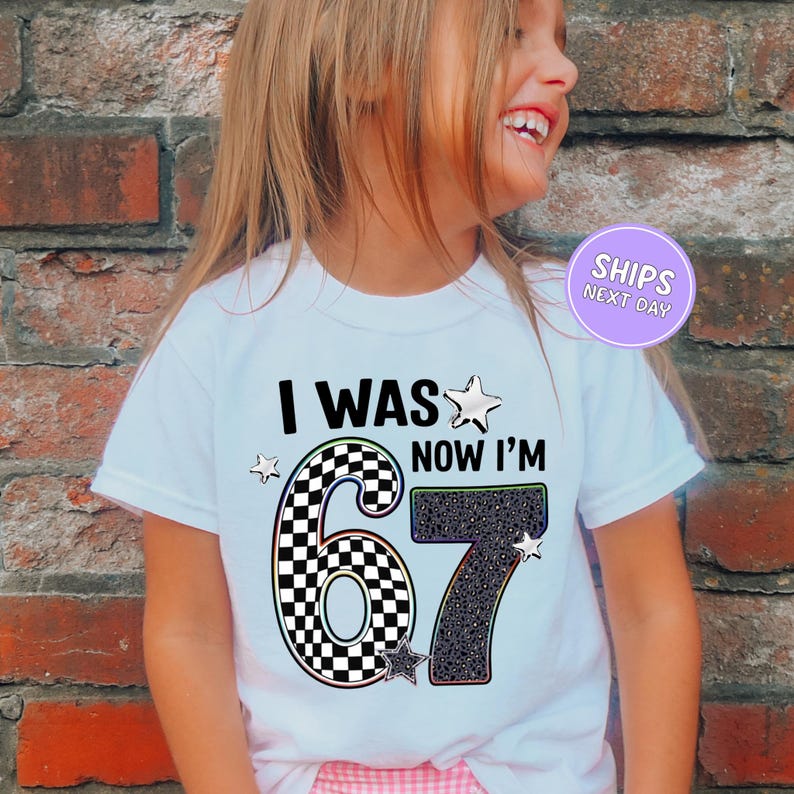 A young girl wearing a white t-shirt with the text "I WAS NOW I'M 67" printed on it, smiling and posing in front of a brick wall.