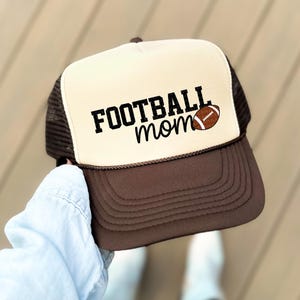 A person is holding a baseball cap with the text "FOOTBALL mom" printed on it.