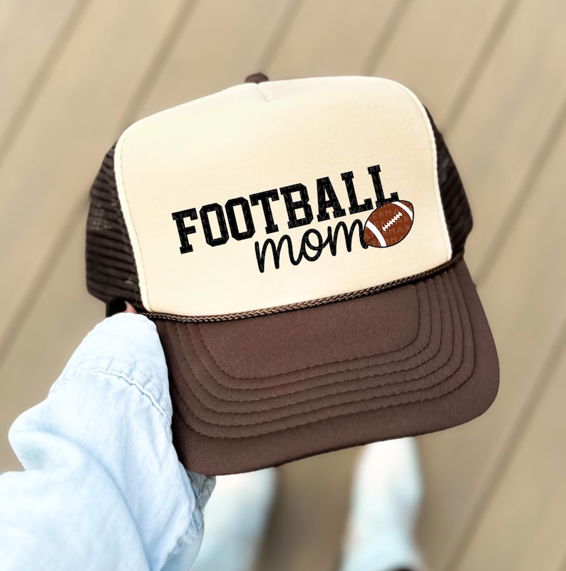 A person is holding a baseball cap with the text "FOOTBALL mom" printed on it.