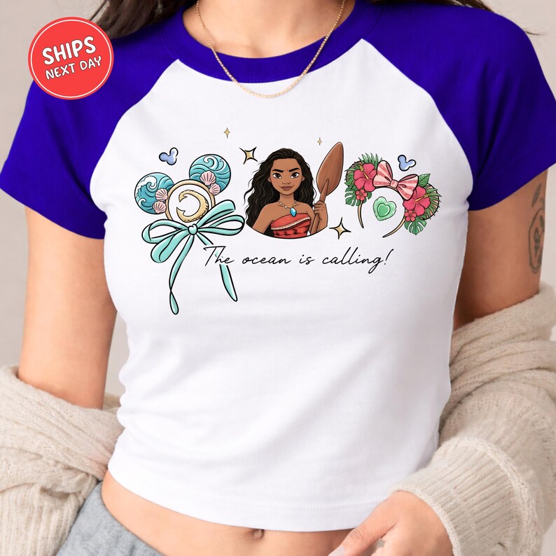 A woman wearing a white and blue t-shirt with a graphic design featuring a cartoon illustration of a young woman surrounded by various ocean-themed elements, including a mermaid-like figure and flowers.