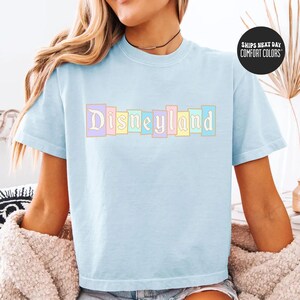 A woman wearing a light blue t-shirt with the word "Disneyland" printed on it, sitting on a bed with a beige blanket.