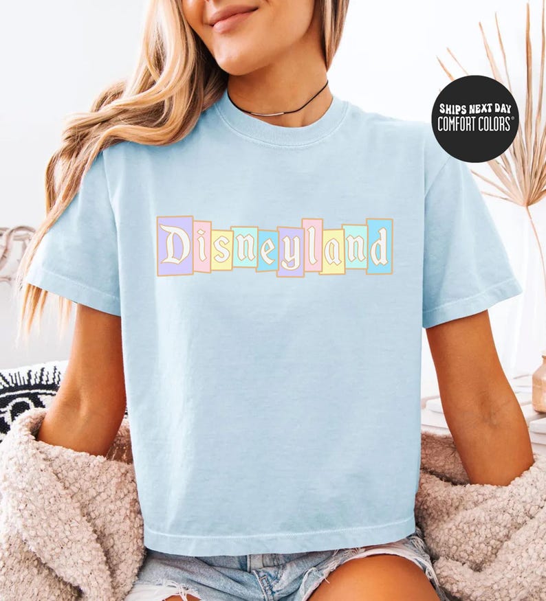 A woman wearing a light blue t-shirt with the word "Disneyland" printed on it, sitting on a bed with a beige blanket.