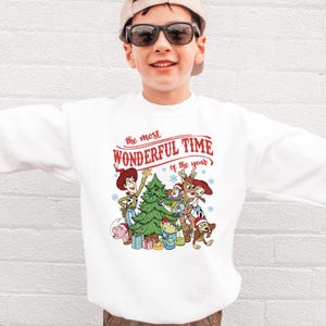 A young boy wearing a white sweatshirt with a Christmas-themed graphic and text printed on it.