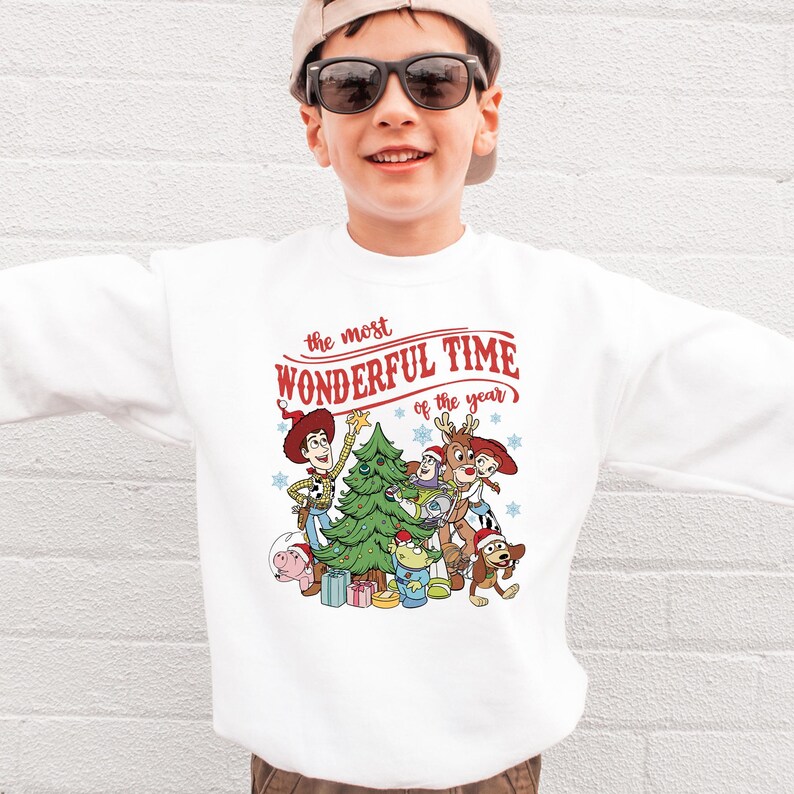 A young boy wearing a white sweatshirt with a Christmas-themed graphic and text printed on it.