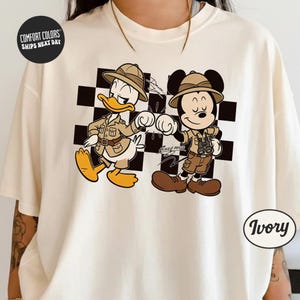 A white t-shirt with a graphic design featuring two cartoon characters, Mickey Mouse and Donald Duck, dressed in safari attire and standing next to each other.