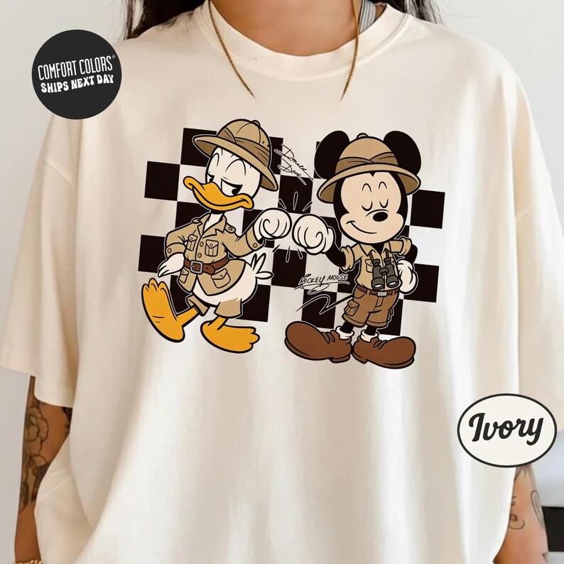 A white t-shirt with a graphic design featuring two cartoon characters, Mickey Mouse and Donald Duck, dressed in safari attire and standing next to each other.