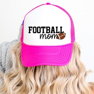 A person with blonde hair wearing a pink and white baseball cap with the text "FOOTBALL mom" printed on it.