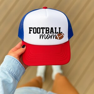 A person is holding a red and white baseball cap with the text "FOOTBALL mom" printed on it.