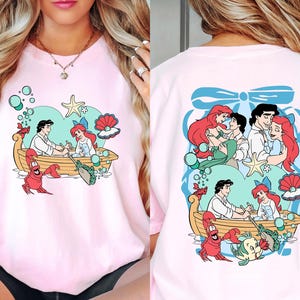 A woman wearing a pink t-shirt with a cartoon design of characters from the Disney film "The Little Mermaid".