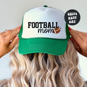 A person is holding up a green and white baseball cap with the text "FOOTBALL mom" printed on it.