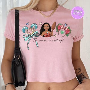 A woman wearing a pink crop top with a graphic design featuring a cartoon character and text that says "The ocean is calling!"