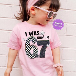A young girl wearing a pink t-shirt with a graphic design and text, standing against a white brick wall.