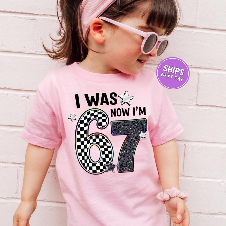 A young girl wearing a pink t-shirt with a graphic design and text, standing against a white brick wall.