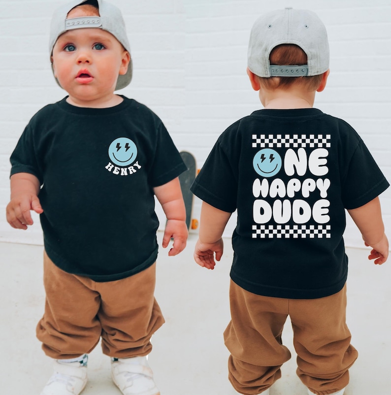 A young child wearing a black t-shirt with a smiley face and the name "HENRY" printed on it, along with a cap and brown pants. The child is standing in front of a white wall.