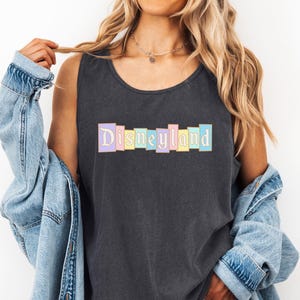 A woman wearing a black tank top with the word "Disneyland" printed on it, along with a denim jacket, stands against a white background.