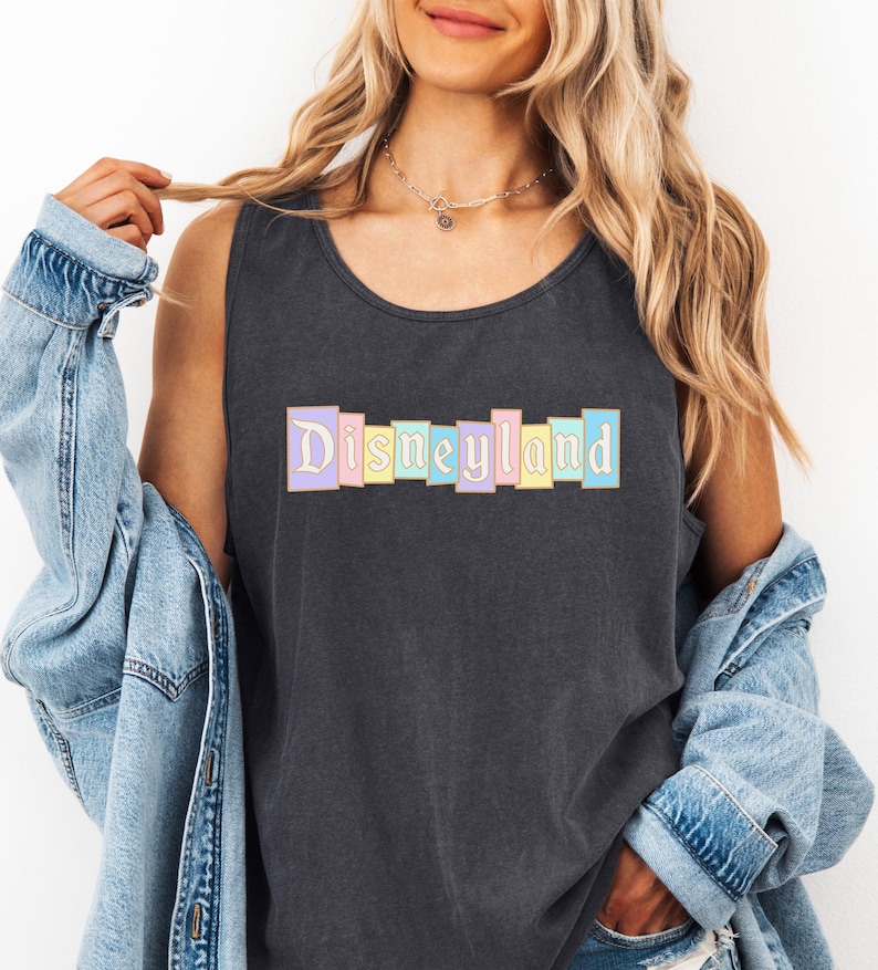 A woman wearing a black tank top with the word "Disneyland" printed on it, along with a denim jacket, stands against a white background.