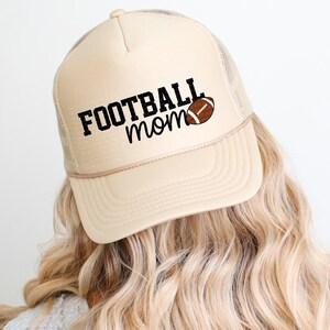 A person with long, wavy blonde hair wearing a beige baseball cap with the text "FOOTBALL mom" printed on it.