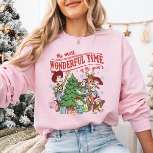 A woman wearing a pink sweatshirt with a Christmas-themed graphic design featuring characters from the Toy Story franchise.