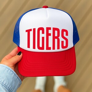 A person's hand holding a red and white baseball cap with the word "TIGERS" printed on it.
