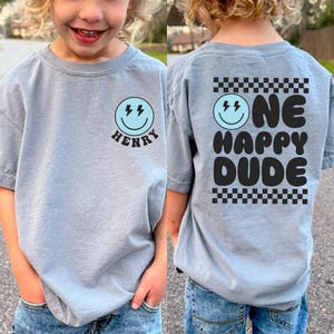 A young child wearing a gray t-shirt with a smiley face and text printed on the back.