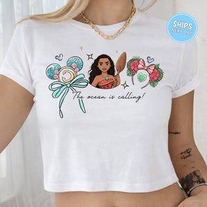A white crop top with a graphic design featuring a woman surrounded by ocean-themed elements, including waves, flowers, and a ribbon.