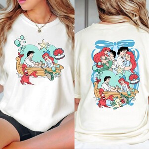 A white t-shirt with a colorful graphic design featuring characters from the Disney animated film "The Little Mermaid".