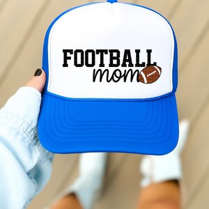 A person's hand holding a blue and white baseball cap with the text "FOOTBALL mom" printed on it.