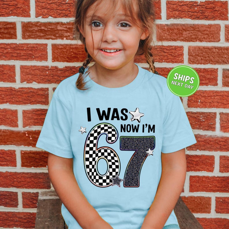 A young girl wearing a light blue t-shirt with a graphic design that says "I WAS NOW I'M 67" and stars.
