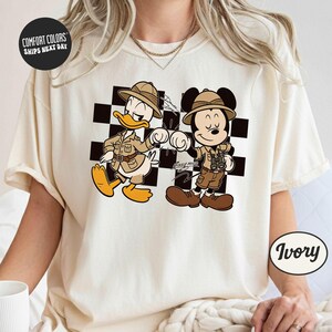 A white t-shirt with a graphic design featuring two cartoon characters, Mickey Mouse and Donald Duck, dressed in safari attire.