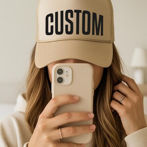 A person is taking a selfie with a tan baseball cap on their head, holding a smartphone in front of their face.