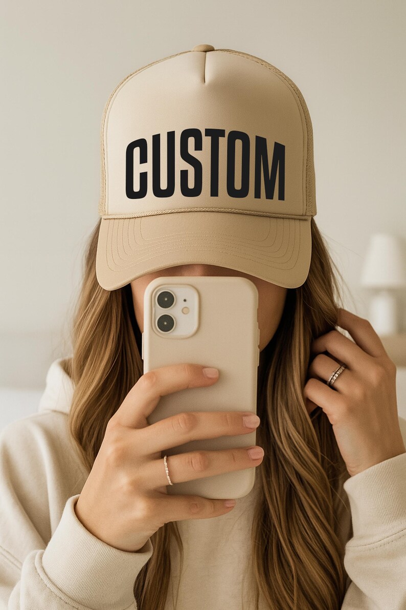 A person is taking a selfie with a tan baseball cap on their head, holding a smartphone in front of their face.