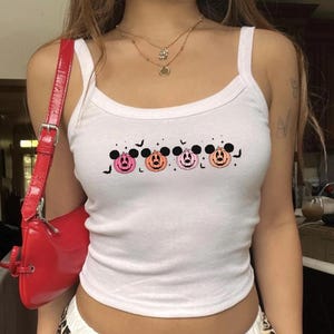 May include: A white sleeveless tank top featuring a graphic print of four pumpkins with black mouse ears, alternating in shades of pink and orange, accented with small black bats and stars. A shiny red shoulder bag with a silver buckle is visible.