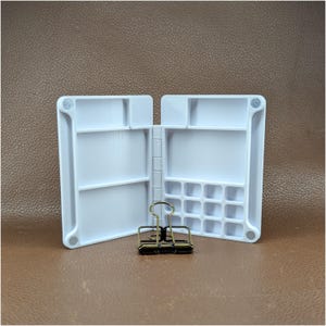 May include: A white plastic folding palette with multiple rectangular and square mixing wells, featuring a hinged design and small circular magnets at the corners. A bronze-colored metal binder clip is positioned in front of the open palette.
