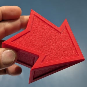 Op de afbeelding: Een felrood pijl-vormig object met een textuur. De pijl heeft een geometrisch ontwerp met een verhoogde rand en een gevouwen gedeelte. De achtergrond is een verloop van blauw en wit.