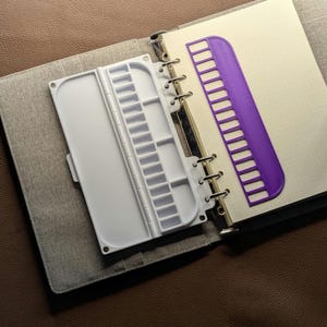 May include: A gray fabric binder containing a white plastic watercolor palette with multiple rectangular mixing wells and a purple plastic thread organizer with twelve slots, both attached to the metal ring mechanism of the notebook.