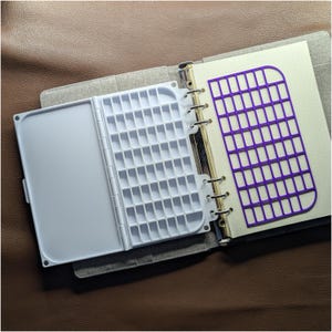 Puede incluir: Una carpeta de tela gris que contiene una paleta de acuarela de plástico blanco con múltiples compartimentos rectangulares y una plantilla de rejilla de plástico morado. La carpeta tiene un mecanismo de anillas metálicas.