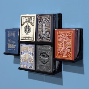 May include: A collection of playing card decks displayed on a black wall shelf. The decks feature various designs, including "Bicycle Standard," "Kansas City," and "Star Wars" themes. The cards are in various colors, including blue, red, and gray.