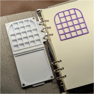 May include: A white plastic watercolor palette with a hinged lid and multiple square mixing wells, attached to the metal rings of a dot-grid notebook. A purple grid-shaped stencil is placed on the notebook page next to the palette.