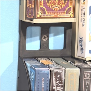 May include: A black plastic wall-mounted display rack for playing card decks. The rack features a central vertical support with two rectangular cutouts and a visible silver screw. Multiple decks of playing cards are arranged in the slots.
