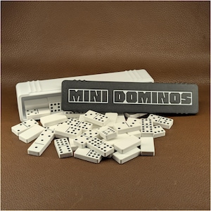 Peut inclure: Un jeu de mini dominos avec un étui blanc et un couvercle noir avec l'inscription "MINI DOMINOS" en lettres blanches. Les dominos sont blancs avec des points noirs, dispersés sur une surface marron.