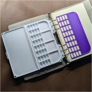 May include: A white plastic folding paint palette with multiple rectangular mixing wells and a separate purple plastic grid stencil insert, both attached to the metal ring binding of a gray fabric-covered notebook with cream-colored pages.