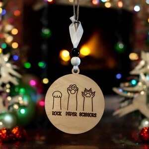 May include: A wooden Christmas bauble featuring a paw print design. The bauble reads "Rock Paper Scissors" with a paw print representing each word.