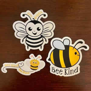 May include: Three bee-themed stickers with different designs and messages. The first sticker features a cartoon bee with black and yellow stripes and the words "Bee Happy". The second sticker features a cartoon bee with black and yellow stripes and the words "Bee Kind". The third sticker features a cartoon bee with black and yellow stripes and the words "Bee Kind" and "Bee Brave".