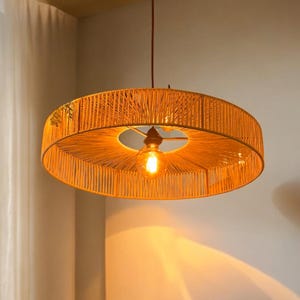 May include: A circular pendant light, woven from a yellow-orange material. The light fitting has a central bulb and is suspended from a brown cord. The woven design gives a textured appearance. The light is suitable for a room.