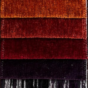 May include: A vertical display of fabric swatches in various colors and textures. The samples range from light neutral tones to deep jewel tones, including shades of beige, brown, gold, red, purple, and teal. The fabrics appear to be plush and textured.